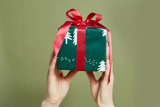 Hands holding green Christmas gift box with white tree pattern and red satin ribbon bow against beige background