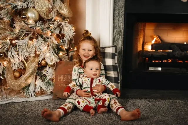Two children in matching Christmas pajamas sitting by decorated tree and fireplace during holiday season