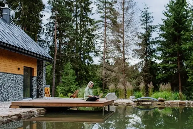 Woman using laptop by natural swimming pool at forest cabin retreat, demonstrating reliable internet connectivity while traveling in remote locations