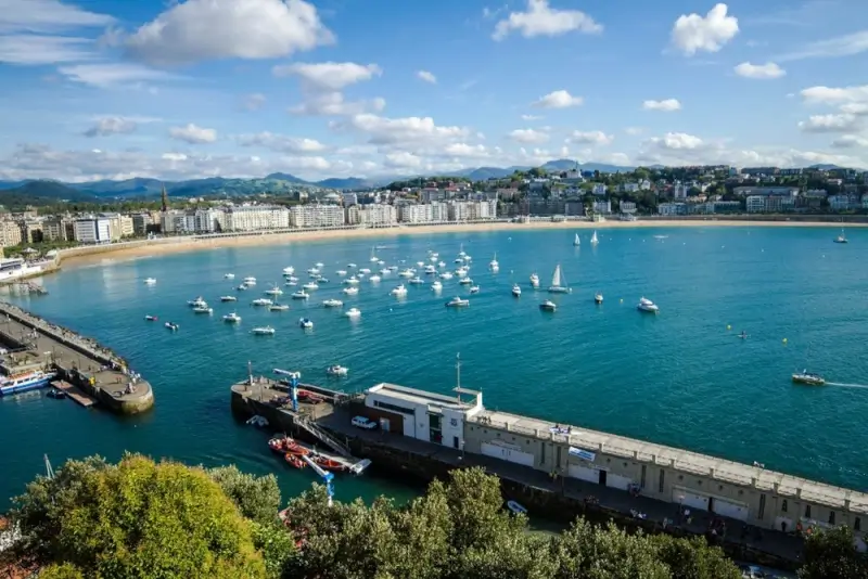 Panoramic aerial view of La Concha Beach in San Sebastian Spain showing the famous crescent-shaped bay with turquoise waters, white sandy beach, Belle Epoque waterfront buildings, and boats anchored in the harbor under blue skies with white clouds