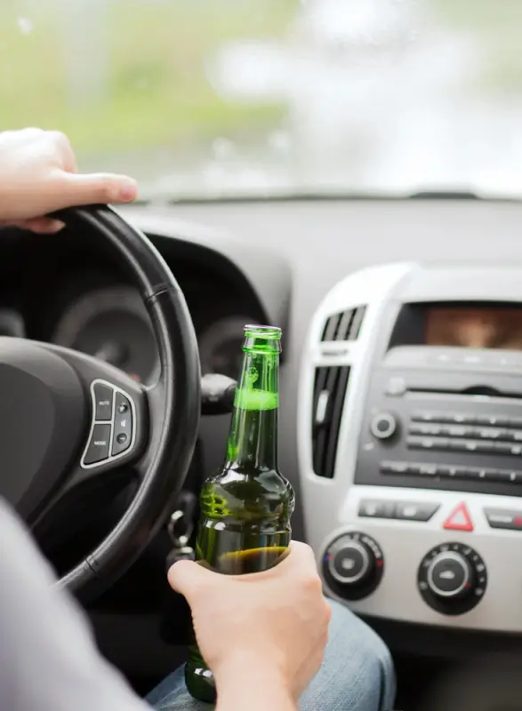 Person holding beer bottle while gripping car steering wheel inside vehicle, illustrating dangerous drunk driving behavior that leads to DUI charges
