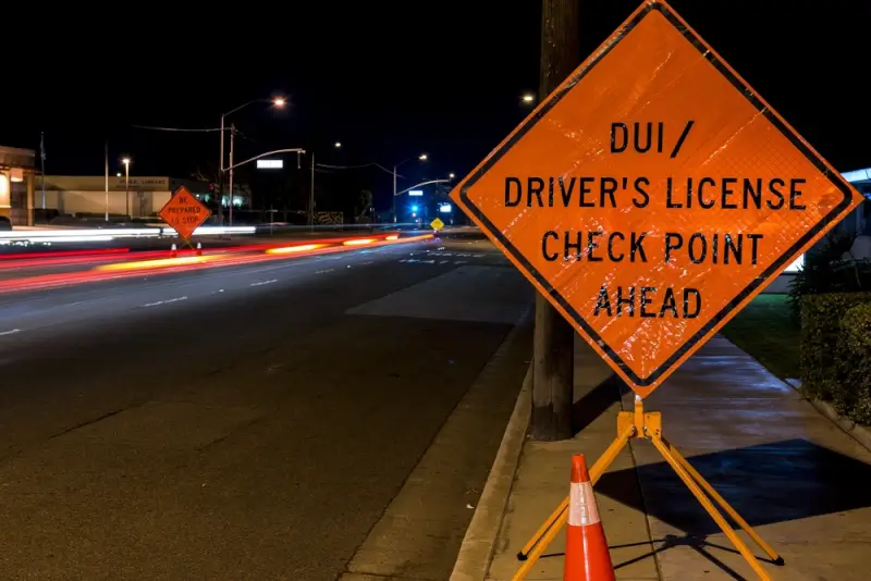 Orange DUI driver's license checkpoint sign on nighttime street with traffic light trails and orange cones, showing police enforcement that can lead to minor DUI arrests
