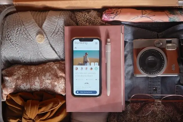 Smartphone displaying travel booking app surrounded by packed travel essentials including clothes, camera, and sunglasses in suitcase