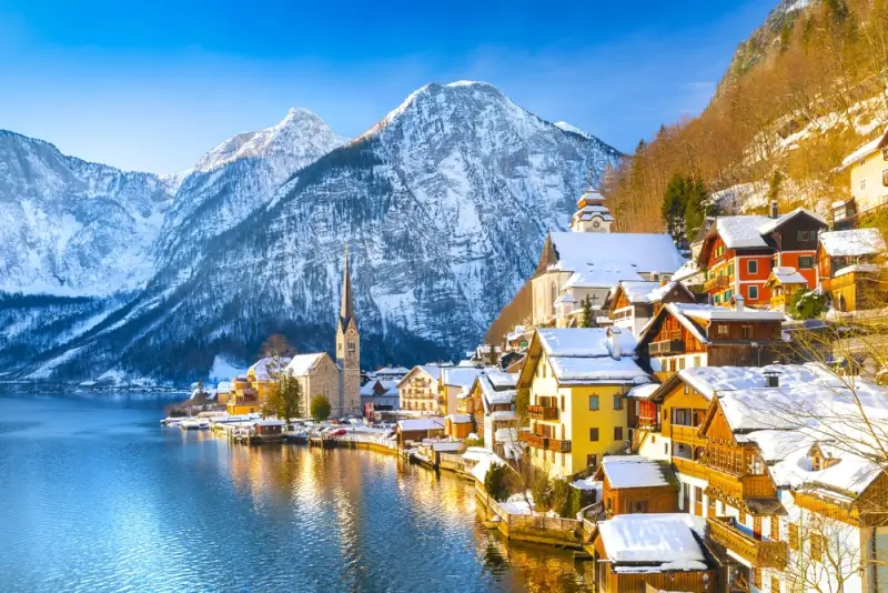 Famous Austrian lakeside village of Hallstatt in winter, with colorful historic buildings, church spire, and snow-covered rooftops reflected in the pristine alpine lake beneath towering mountains