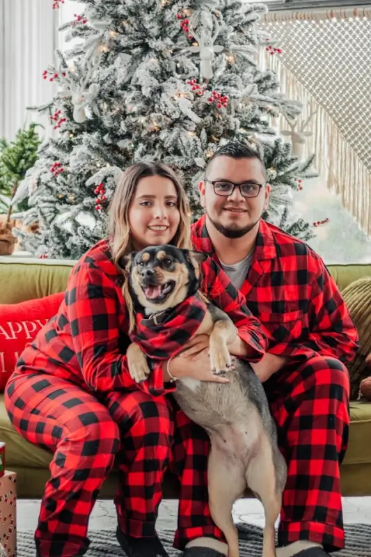 Couple in matching red buffalo plaid Christmas pajamas with dog by flocked Christmas tree in living room