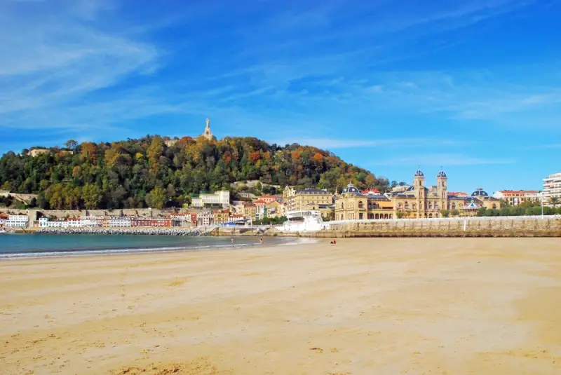 La Concha Beach in San Sebastian with golden sand and calm waters, showing the Old Town colorful buildings, Monte Urgull hill with autumn foliage, and the iconic City Hall building under bright blue skies