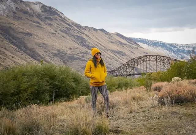Woman wearing striped cotton leggings and yellow sweater standing in mountain landscape with bridge in background