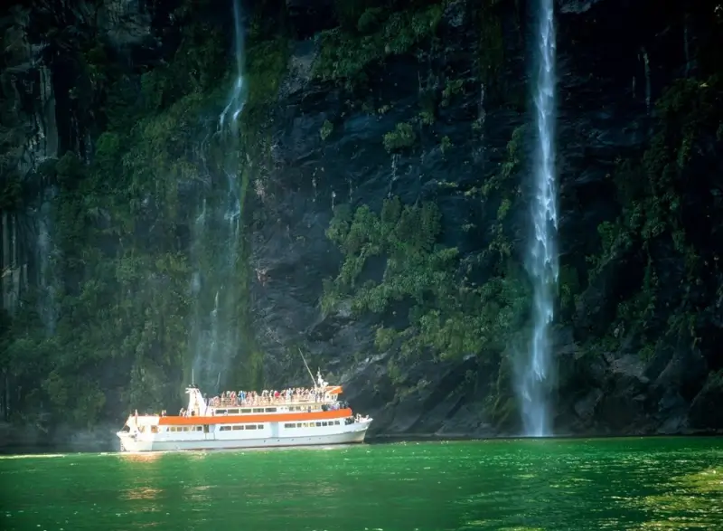 Small river cruise ship with passengers on deck sailing past dramatic waterfall cascading down moss-covered cliff face