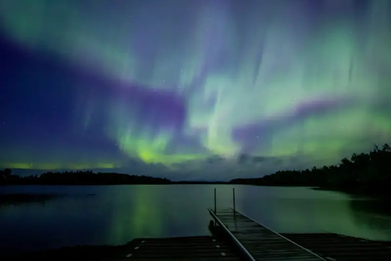 Northern lights aurora borealis dancing over Crane Lake with green and purple colors reflecting on water from wooden dock