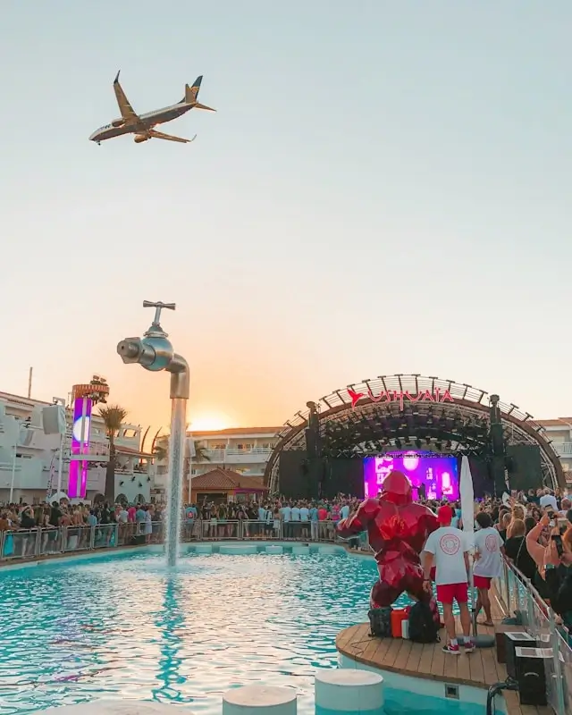 Ibiza beach club pool party at sunset with DJ stage and airplane flying overhead