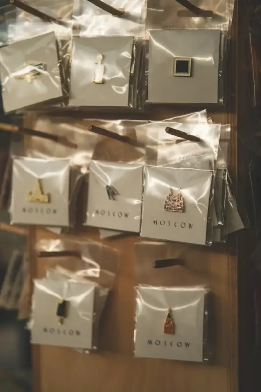 Display of enamel pins in clear plastic packaging arranged in a wooden rack. Each package is labeled 'MOSCOW' and contains a single pin with different designs including landmarks, architectural elements, and cultural symbols. The packages are organized in multiple rows, creating a retail merchandise display