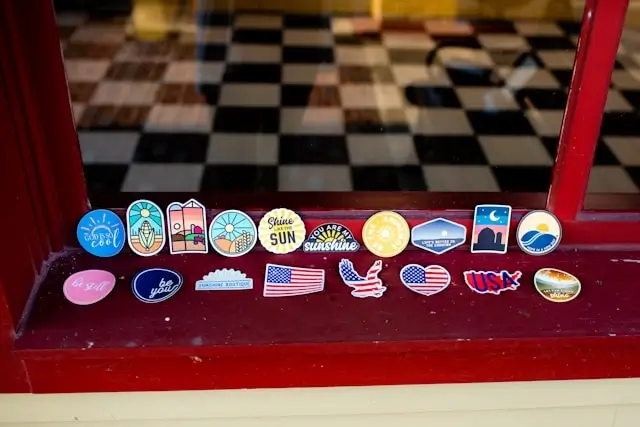 Collection of enamel pins displayed on the red metal edge of a table with a black and white checkered surface. The pins include various designs such as American flags, travel destinations, landmarks, logos, and text-based pins arranged in two rows along the table's border