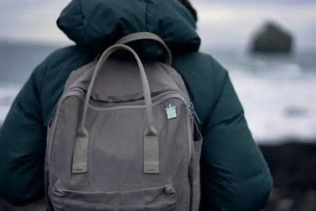 Person wearing a dark green puffy jacket and gray backpack with a small enamel pin attached, standing on a beach with ocean waves and a large rock formation visible in the blurred background