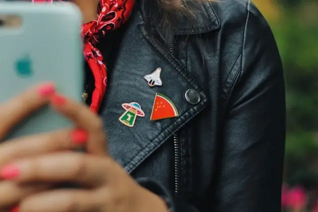 Close-up of a person wearing a black leather jacket with three enamel pins attached to the lapel: a white paper airplane, a red and green mushroom, and an orange watermelon slice. A red bandana is visible around their neck, and blurred hands holding a phone are in the foreground