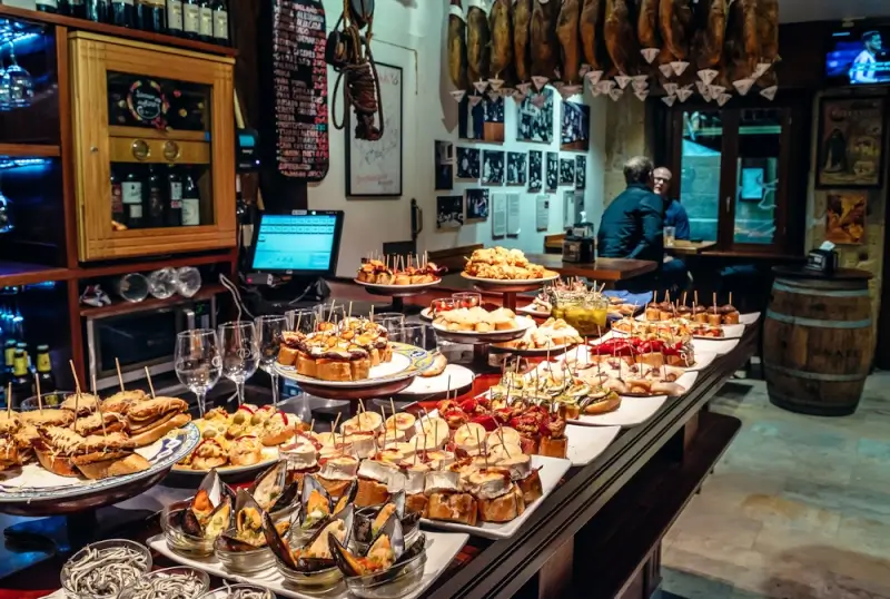 Traditional pintxos bar counter in San Sebastian Old Town displaying colorful Basque tapas on toothpicks with wine glasses, cured ham hanging overhead, and bartender serving customers in authentic Spanish tavern