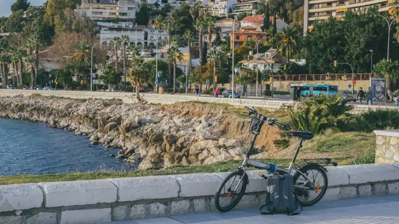 Folding bicycle parked along Mediterranean coastal promenade with rocky shoreline and hillside town in background during golden hour