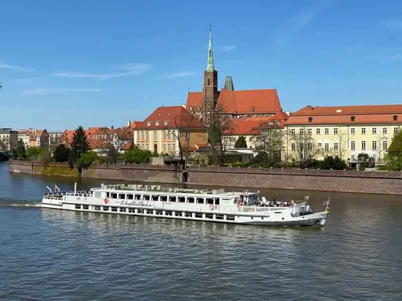 White river cruise ship sailing past historic European city with red-roofed cathedral and traditional architecture along riverbank