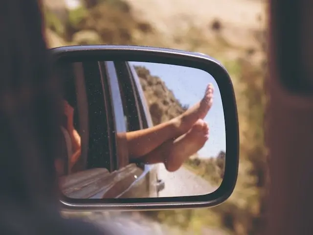 Person's feet hanging out car window reflected in side mirror during budget road trip getaway