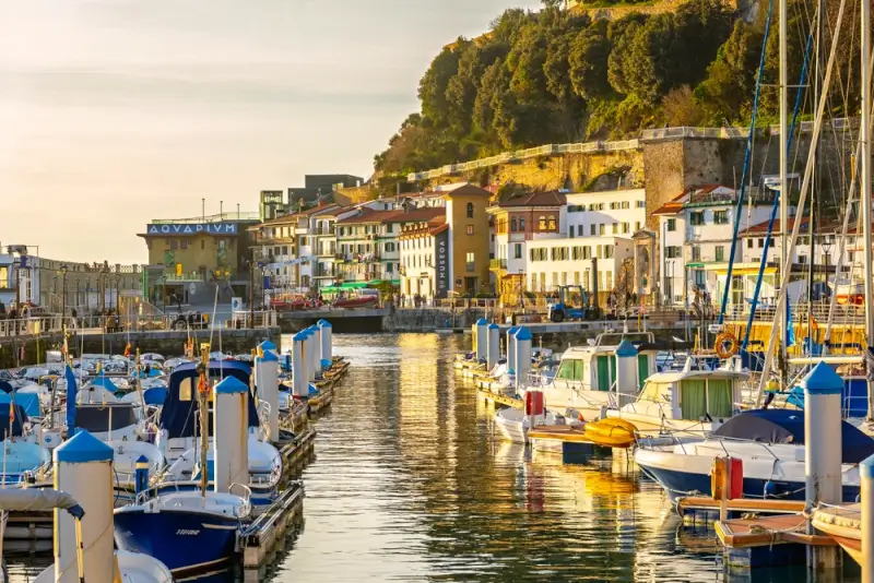 San Sebastian harbor and marina at golden hour with fishing boats, sailboats, and colorful waterfront buildings of the Old Town, including the iconic Aquarium building, perfect for cycling tours ending with waterfront pintxos
