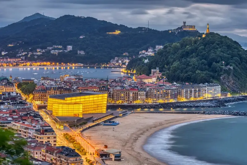 San Sebastian panoramic twilight view showing Monte Igueldo and Monte Urgull hills, La Concha Bay, Zurriola Beach, the golden Kursaal Congress Centre, and illuminated Old Town - spectacular cycling route scenery at dusk