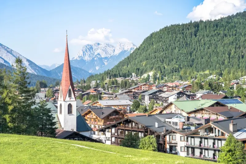 Austrian Alpine village, Seefeld, in summer with distinctive red-spired church, traditional wooden chalets and modern buildings nestled in green valley surrounded by forested mountains and snow-capped peaks