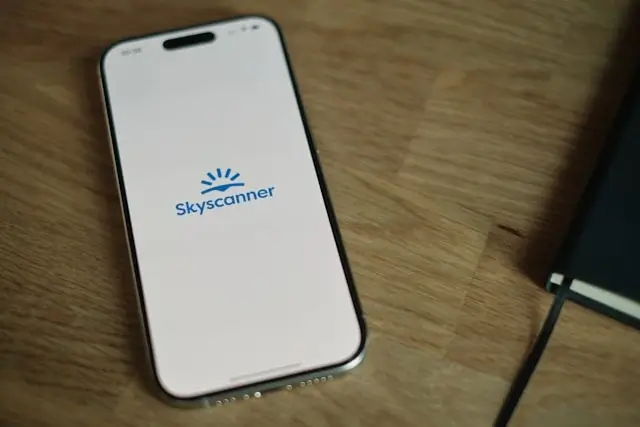 Smartphone displaying Skyscanner flight booking app on wooden desk with notebook for travel planning