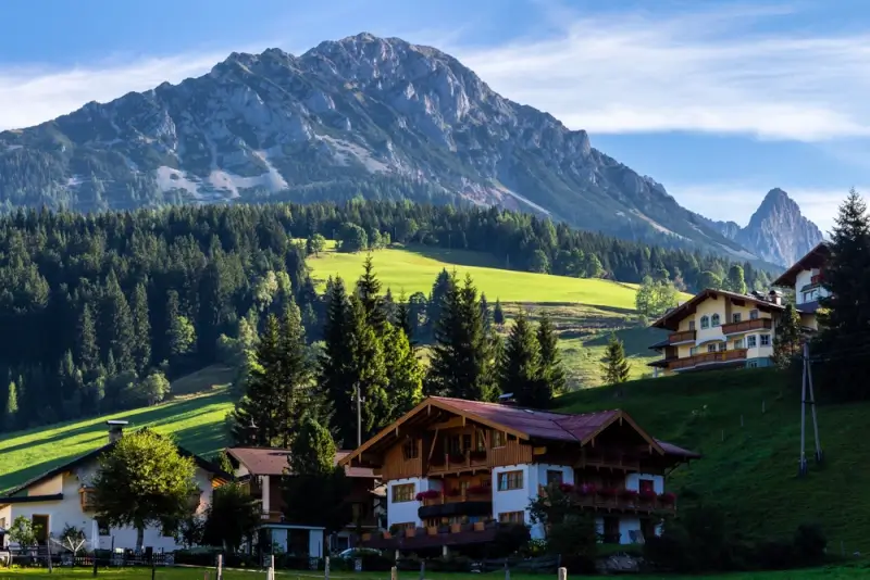 Traditional Austrian Alpine chalets in Filzmoos with flower-filled balconies nestled on green hillsides beneath towering limestone peaks and dramatic rocky spires in summer sunlight