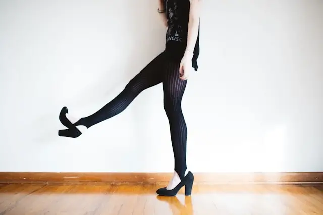 Woman in black ribbed cotton leggings and heels doing dynamic pose against white wall on wooden floor