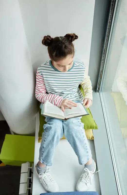 Young girl focused on reading a book while sitting by a sunny window at home