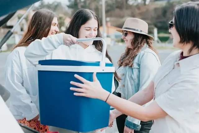 Group of friends gathered around a blue portable car cooler during a road trip, with one person opening the lid while others look on excitedly near their parked vehicle