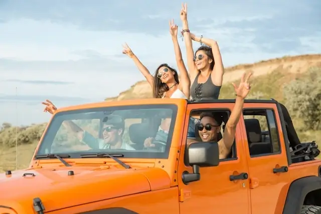 Happy friends celebrating in an orange off-road vehicle during a road trip adventure, with two people standing through the sunroof with arms raised against a coastal landscape