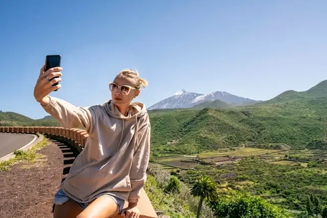 Female traveler taking a selfie with smartphone against scenic mountain landscape and lush green valley