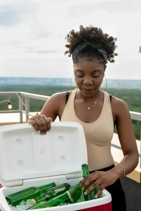 Woman opening a white portable cooler filled with chilled beverages and ice during an outdoor adventure with scenic landscape in the background