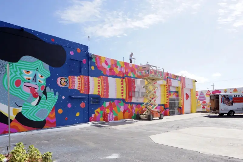 Artists painting colorful street art murals on warehouse walls in Miami's Wynwood district, featuring vibrant geometric patterns and a green face character against a blue starry background
