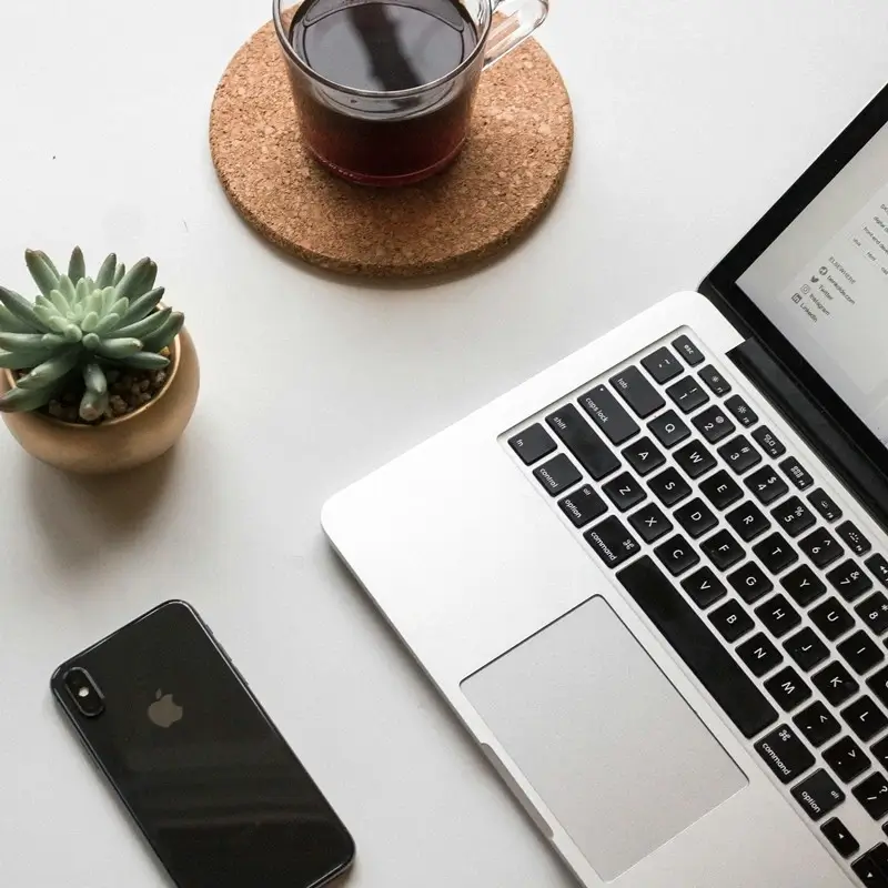 Minimalist content creator workspace flatlay featuring laptop, smartphone, coffee cup on cork coaster, and small succulent plant on white desk