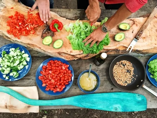 Overhead view of hands chopping fresh vegetables on a wooden cutting board surrounded by colorful bowls of diced tomatoes, cucumbers, leafy greens, and cooking ingredients