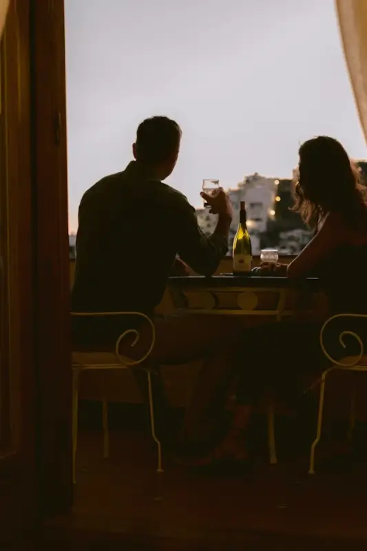 Couple sharing wine at sunset on balcony overlooking Mediterranean cityscape, silhouetted against evening sky