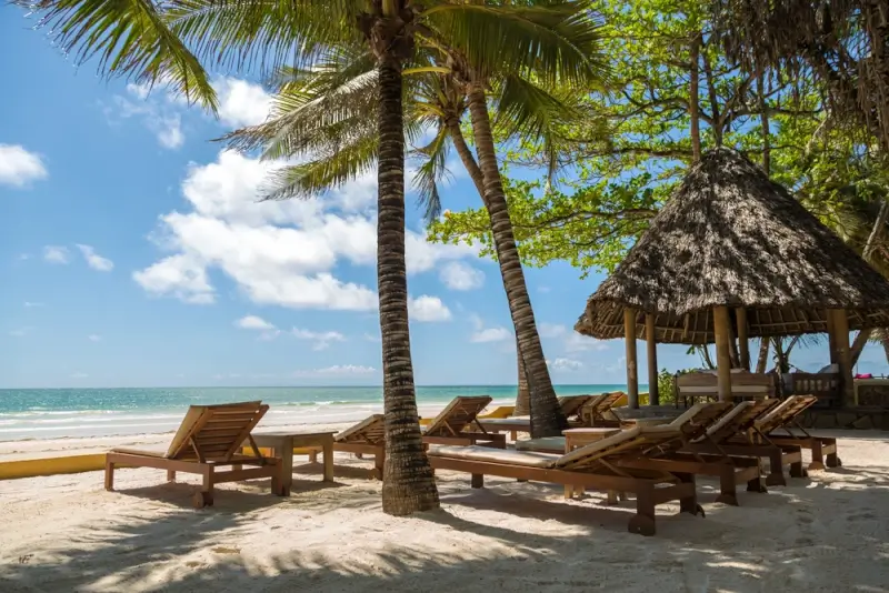 Wooden beach loungers and thatched roof shelter on white sand Diani Beach with palm trees and turquoise Indian Ocean waters in Kenya