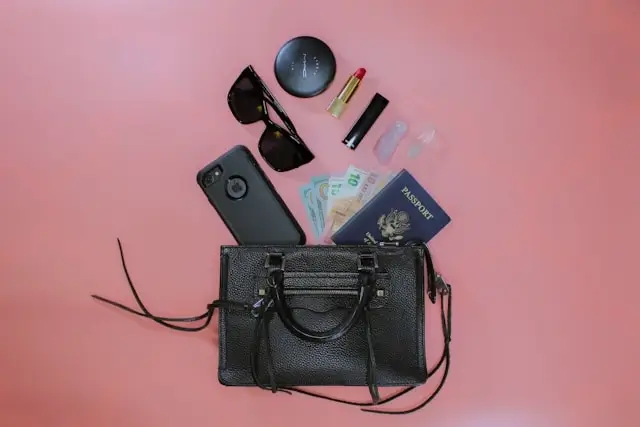 Essential items for travel laid out on pink background including black leather handbag, passport with currency, sunglasses, smartphone, cosmetics, and travel accessories