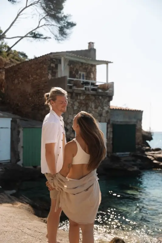Young couple enjoying a sunny moment by the Mediterranean coast with rustic stone buildings and calm blue water in the background