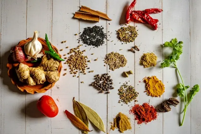 Assortment of aromatic Indian spices including turmeric, cumin, coriander, and dried chilies arranged on white wooden surface with fresh ginger, garlic, and cilantro