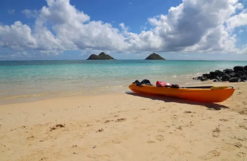 Orange kayak on sandy beach with twin Mokulua Islands visible across turquoise waters under a cloudy blue sky in Kailua, Oahu