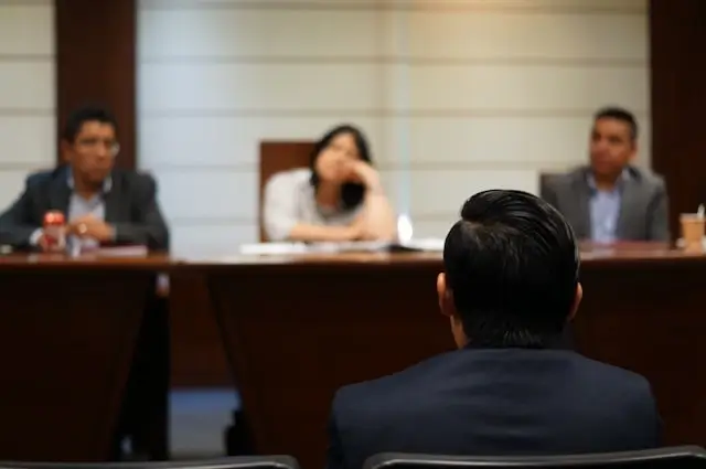 Person seated at a conference table facing three panel members during a formal discussion or legal meeting.