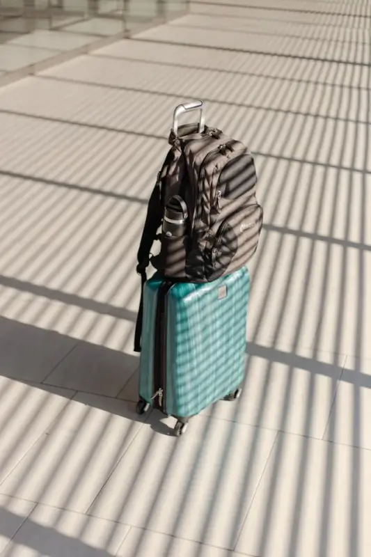 Turquoise rolling suitcase with gray backpack attached on top, casting shadows on tiled floor at airport or station