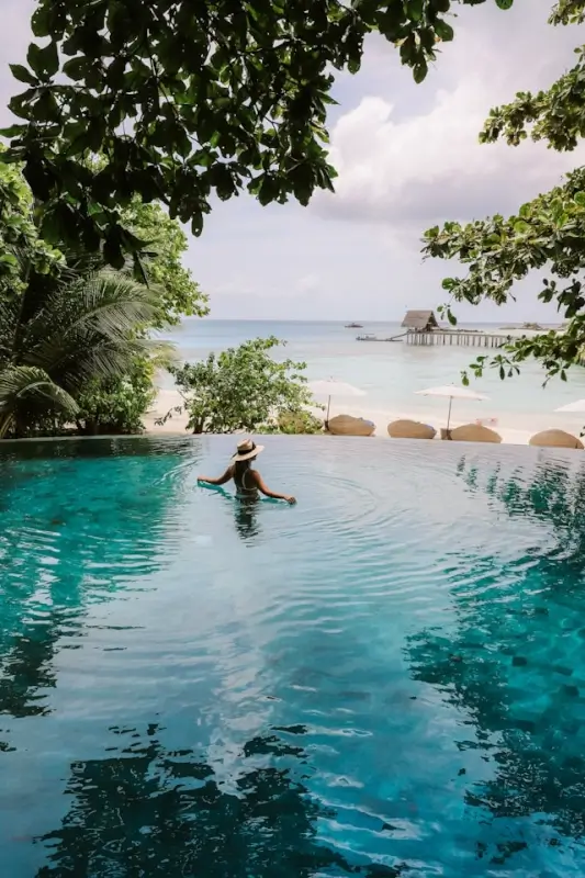 Woman in sun hat relaxing in tropical infinity pool overlooking turquoise ocean at cheap luxury destination resort