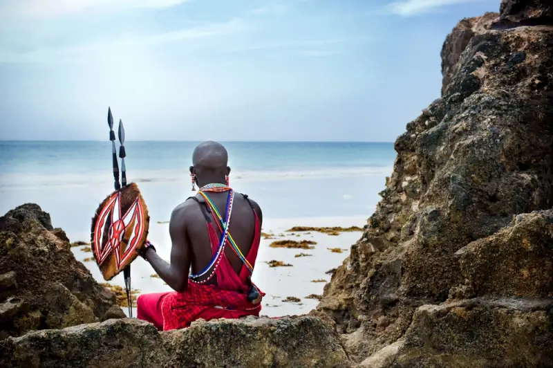 Maasai warrior in traditional red shuka and beaded jewelry sitting on coastal rocks overlooking Indian Ocean beach in Kenya