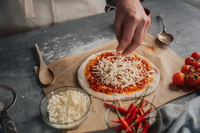 Hand sprinkling mozzarella cheese on homemade pizza dough with tomato sauce, surrounded by fresh ingredients and wooden utensils on kitchen counter