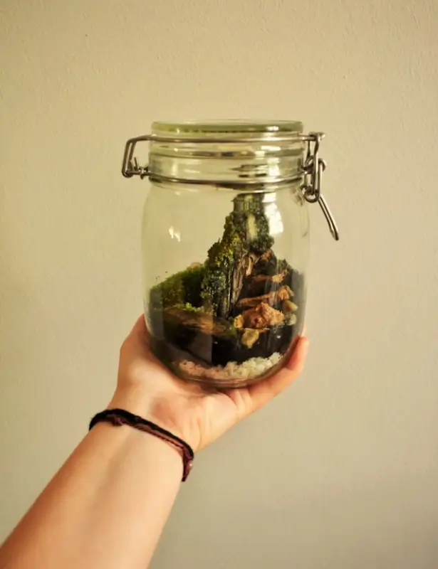 Hand holding a glass clamp jar containing a miniature terrarium with moss, small rocks, and natural elements collected as travel keepsakes against a beige background