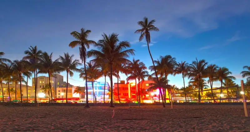 Iconic Art Deco hotels illuminated with colorful neon lights along South Beach at dusk, with palm trees silhouetted against the evening sky and sandy beach in the foreground
