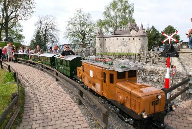 Rideable miniature steam train with passengers passing Swiss castle replica at Swiss Vapeur Parc in Le Bouveret